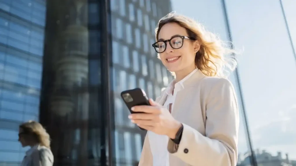 Ecommerce: How to build an online store with technology, user experience and artificial intelligence Mujer sonriente usando un teléfono inteligente frente a un edificio moderno, simbolizando la confianza y la experiencia del usuario en el comercio electrónico.